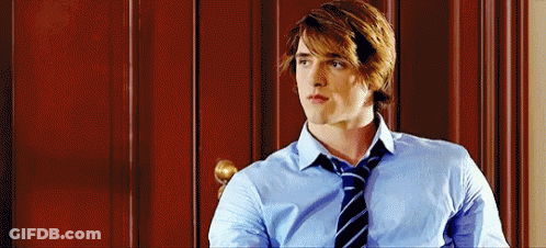 A Man In A Blue Shirt And Tie Is Sitting In Front Of A Wooden Wall . GIF