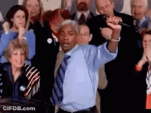A Man In A Blue Shirt And Tie Is Standing In Front Of A Crowd Of People Holding A Microphone . GIF