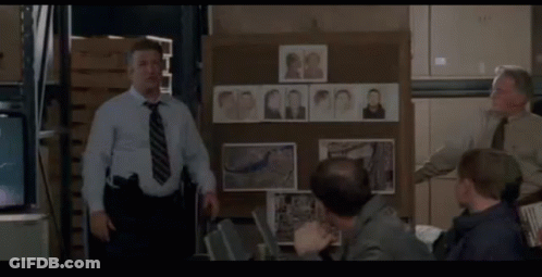 A Man In A Blue Shirt And Tie Stands In Front Of A Board With Pictures Of People On It GIF