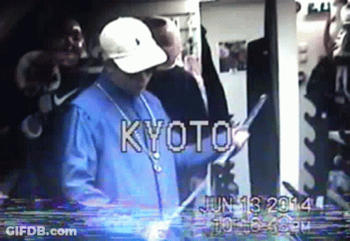 A Man In A Blue Shirt And White Hat Is Standing In A Room With The Word Kyoto On The Screen GIF