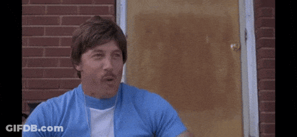 A Man In A Blue Shirt And White Shirt Is Sitting In Front Of A Brick Building . GIF