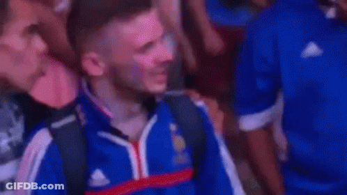 A Man In A Blue Shirt Is Covering His Face With His Hand While Standing In A Crowd . GIF
