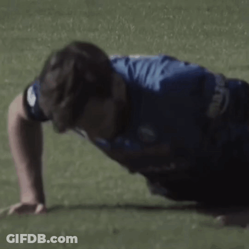 A Man In A Blue Shirt Is Doing Push Ups On A Green Field . GIF