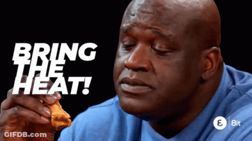 A Man In A Blue Shirt Is Eating A Chicken Wing With The Words " Bring The Heat " Behind Him GIF