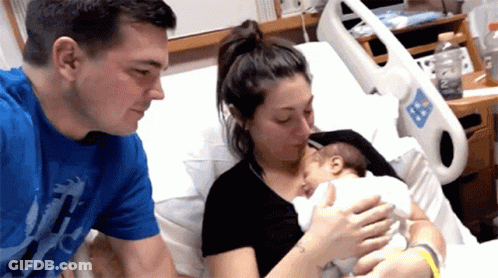 A Man In A Blue Shirt Is Holding A Baby In A Hospital Bed GIF