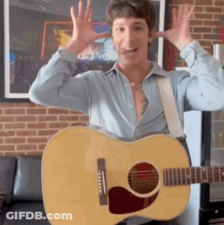 A Man In A Blue Shirt Is Holding A Guitar In Front Of A Brick Wall GIF