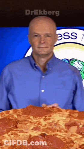 A Man In A Blue Shirt Is Holding A Large Pepperoni Pizza In Front Of A Sign That Says Ess GIF