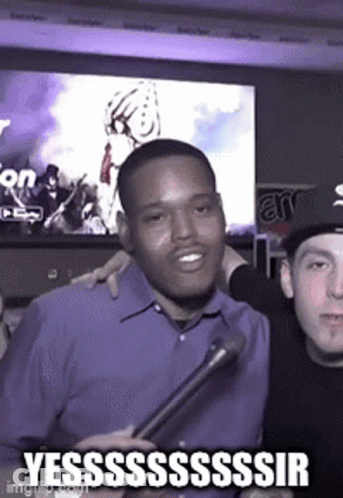 A Man In A Blue Shirt Is Holding A Microphone And Smiling While Another Man Looks On . GIF