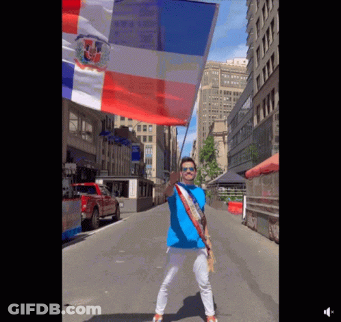 A Man In A Blue Shirt Is Holding A Red White And Blue Flag On A City Street GIF