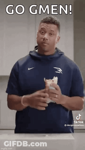 A Man In A Blue Shirt Is Holding A Sandwich In His Hands And Making A Funny Face . GIF
