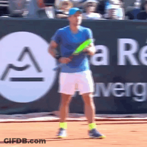 A Man In A Blue Shirt Is Holding A Tennis Racquet In Front Of A Sign That Says A GIF