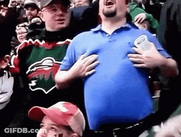 A Man In A Blue Shirt Is Holding His Chest While Standing In A Crowd Of People . GIF