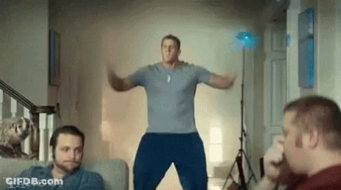 A Man In A Blue Shirt Is Jumping In The Air In A Living Room With Two Other Men . GIF