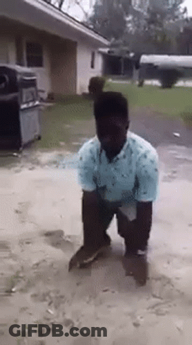 A Man In A Blue Shirt Is Kneeling Down In The Dirt In Front Of A House . GIF