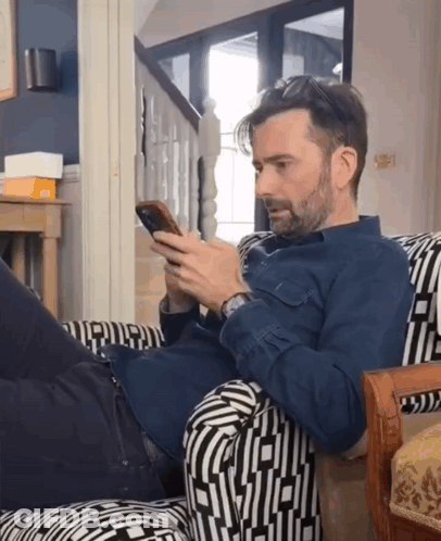 A Man In A Blue Shirt Is Laying On A Chair Looking At His Phone GIF