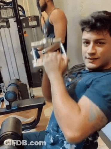 A Man In A Blue Shirt Is Lifting A Dumbbell In A Gym . GIF