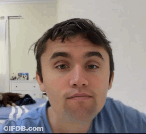 A Man In A Blue Shirt Is Making A Funny Face While Sitting On A Bed . GIF