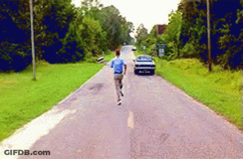A Man In A Blue Shirt Is Running Down A Road With A Car In The Background GIF