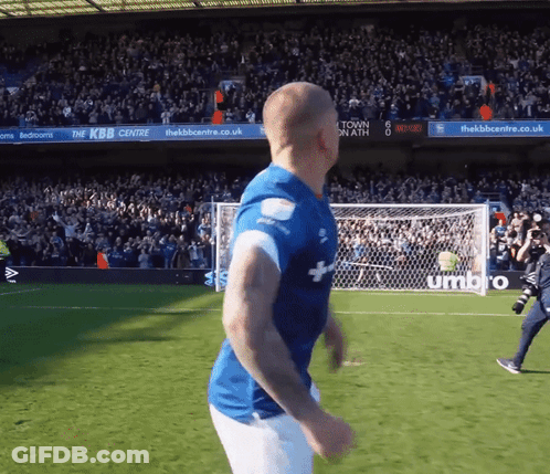 A Man In A Blue Shirt Is Running On A Soccer Field In Front Of A Sign That Says Umbro GIF