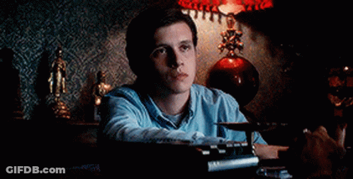 A Man In A Blue Shirt Is Sitting At A Table With A Typewriter And A Lamp In The Background . GIF