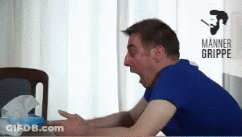 A Man In A Blue Shirt Is Sitting At A Table With His Mouth Open In Front Of A Sign That Says Manner Grippe GIF