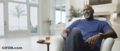 A Man In A Blue Shirt Is Sitting In A Chair In A Living Room With A Glass Of Whiskey On A Table . GIF