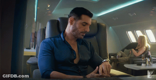 A Man In A Blue Shirt Is Sitting In A Chair Looking At His Watch GIF