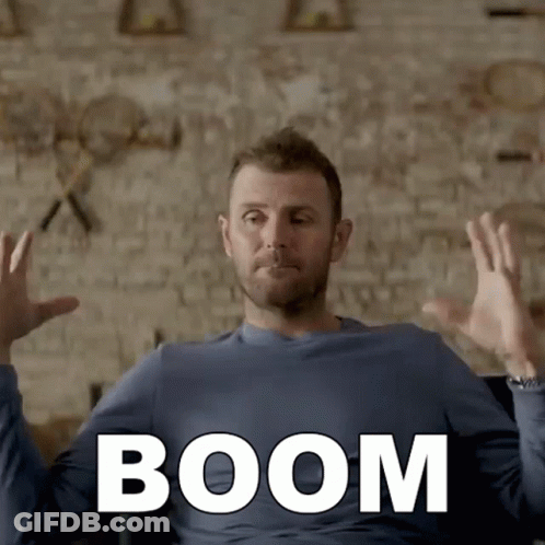 A Man In A Blue Shirt Is Sitting In Front Of A Brick Wall With The Word Boom Written On It GIF