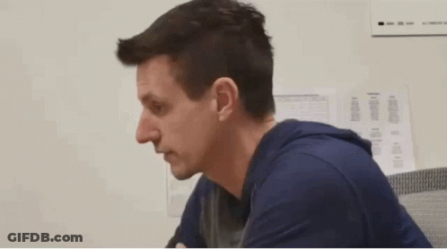 A Man In A Blue Shirt Is Sitting In Front Of A White Wall With Papers On It GIF