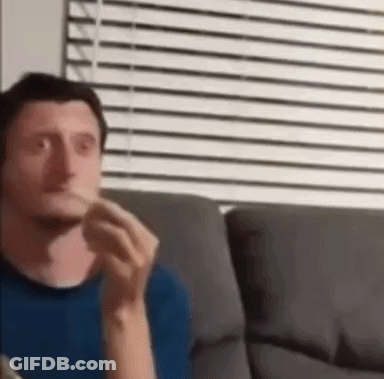 A Man In A Blue Shirt Is Sitting On A Couch Eating A Sandwich . GIF