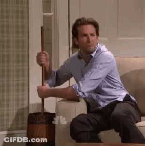 A Man In A Blue Shirt Is Sitting On A Couch Holding A Mop . GIF