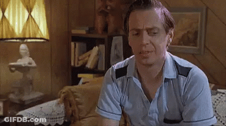 A Man In A Blue Shirt Is Sitting On A Couch In A Living Room With A Lamp In The Background . GIF