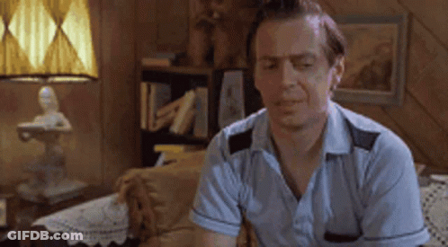 A Man In A Blue Shirt Is Sitting On A Couch In A Living Room With A Lamp . GIF