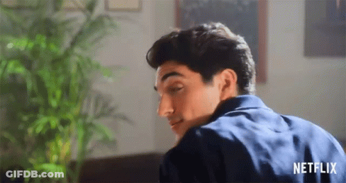 A Man In A Blue Shirt Is Sitting On A Couch In A Living Room With A Plant In The Background . GIF