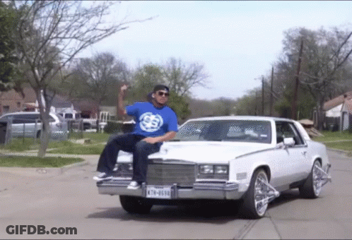 A Man In A Blue Shirt Is Sitting On The Hood Of A White Car With A License Plate That Says K78-6030 GIF