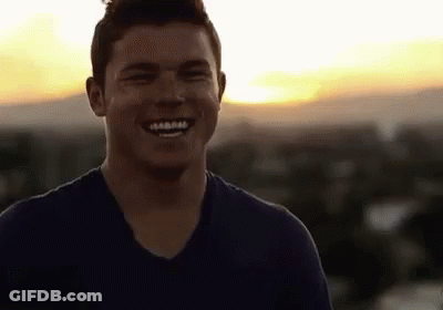 A Man In A Blue Shirt Is Smiling At The Camera While Standing On Top Of A Building At Sunset . GIF