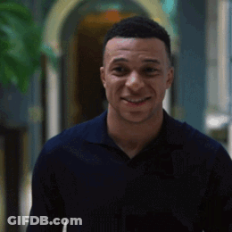 A Man In A Blue Shirt Is Smiling While Standing In A Hallway . GIF