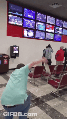 A Man In A Blue Shirt Is Squatting Down In Front Of A Wall Of Televisions Including One That Says ' Nfl ' GIF