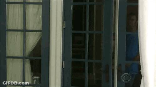 A Man In A Blue Shirt Is Standing In A Doorway Looking Out . GIF