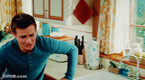 A Man In A Blue Shirt Is Standing In A Kitchen Next To A Sink And Window . GIF
