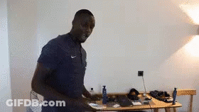 A Man In A Blue Shirt Is Standing In A Room Next To A Desk . GIF