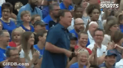 A Man In A Blue Shirt Is Standing In Front Of A Crowd Of People With Spectrum Sports Written On The Bottom Right GIF