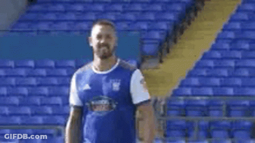 A Man In A Blue Shirt Is Standing In Front Of A Stadium . GIF