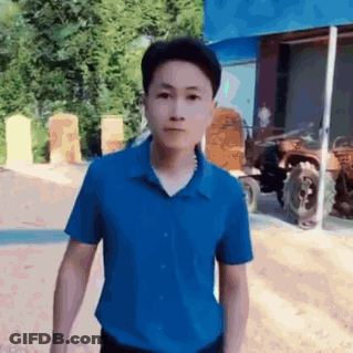 A Man In A Blue Shirt Is Standing In Front Of A Tractor And Looking At The Camera . GIF