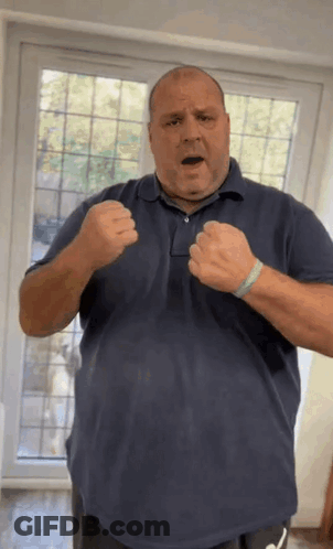 A Man In A Blue Shirt Is Standing In Front Of A Window With His Fists Up . GIF