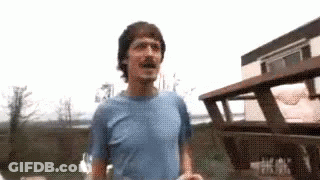 A Man In A Blue Shirt Is Standing In Front Of A Wooden Building . GIF