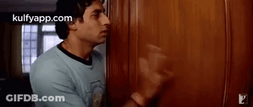 A Man In A Blue Shirt Is Standing In Front Of A Wooden Door In A Room . GIF