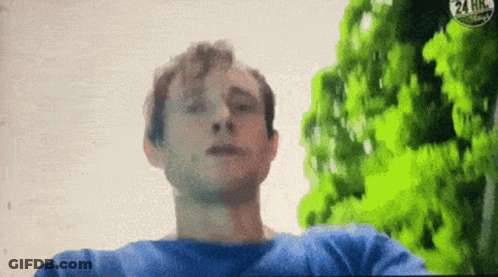 A Man In A Blue Shirt Is Standing In Front Of Trees And A 24 Hr Sign GIF