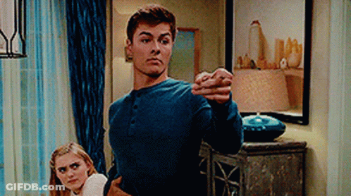 A Man In A Blue Shirt Is Standing Next To A Woman In A Living Room And Pointing At The Camera . GIF
