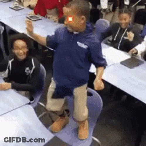 A Man In A Blue Shirt Is Standing On A Chair In Front Of A Group Of People GIF